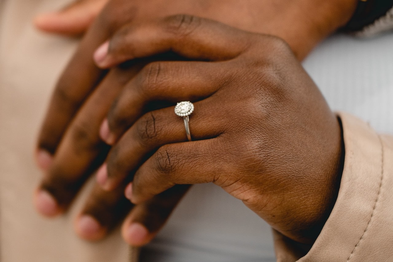 A person’s hand draped over another’s, wearing a halo, oval cut engagement ring