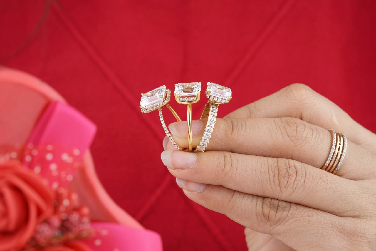 A hand holding three emerald cut engagement rings against a red background