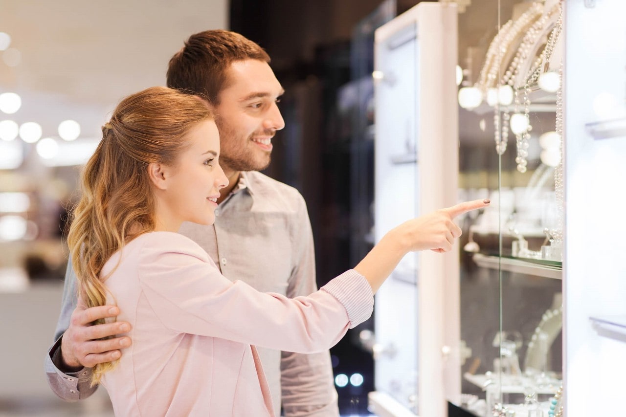 A red-headed woman points out a pearl necklace to her husband while they browse fashion jewelry at a local jewelry store