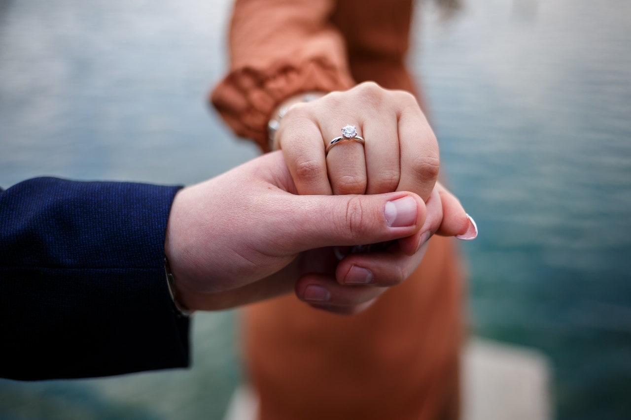 A man holds his fiance’s hand at a lake, showing off her solitaire engagement ring